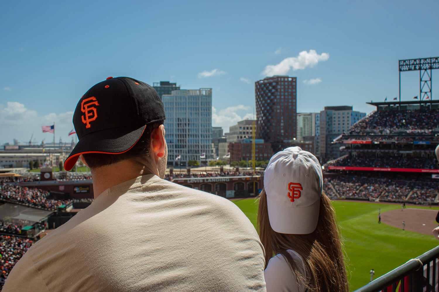 Two people wearing baseball caps with "SF" logos watch a baseball game in a sunny stadium, symbolizing new beginnings. Buildings and a lively crowd are visible in the background, setting the stage for this exciting new chapter.