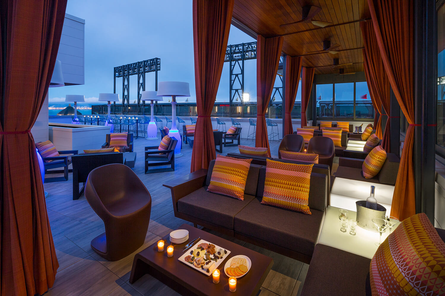 The romantic outdoor lounge area at night features cozy sofas, plush cushions, and a table adorned with candles, snacks, and drinks. With a February chill in the air and a view of the stadium, it's the perfect setting for a serene Valentine's evening.