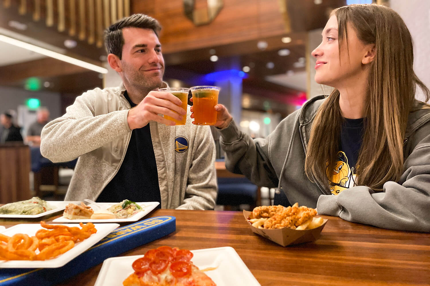 Two people sitting indoors clinking glasses over a table filled with various foods, including pizza and curly fries—a perfect setup for sharing romantic gifts during Valentine's Month.