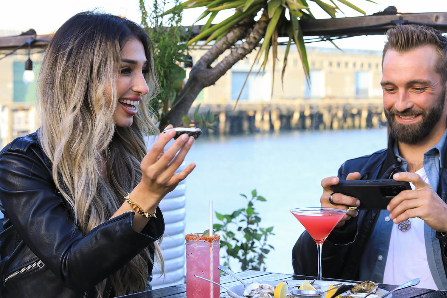 Two people sit at an outdoor table by the water, savoring drinks and oysters. It's Valentine's Month, with one person holding a phone to capture the moment while the other holds an oyster, joyfully immersed in this romantic celebration by the sea.