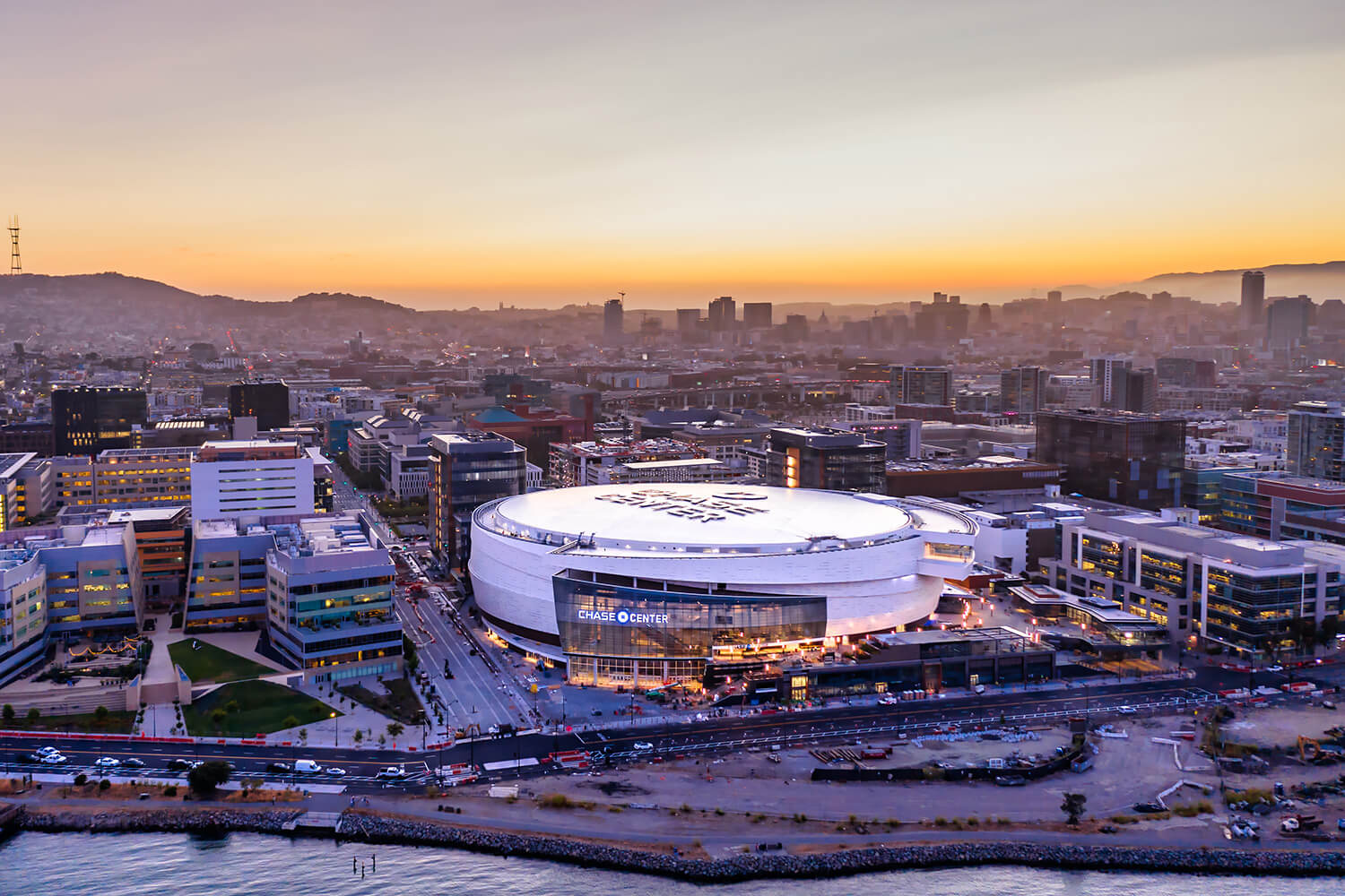 Aerial view of an urban landscape at sunset in February, featuring a large circular arena labeled "Chase Center" surrounded by buildings and roads.