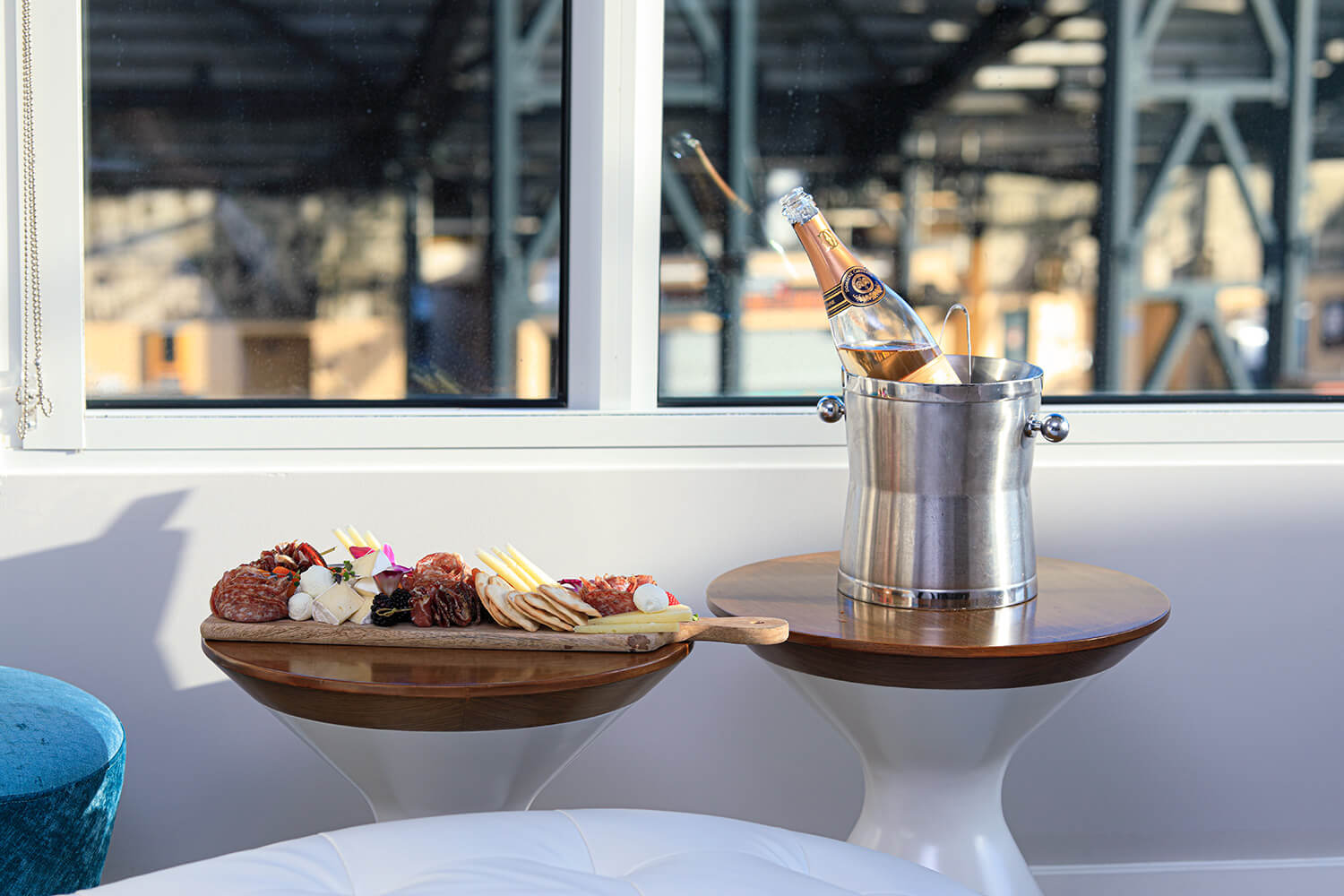 A charcuterie board with meats, cheeses, and fruit sits on a table beside a champagne bottle in an ice bucket by the window, setting the perfect romantic scene for Valentine's Month.