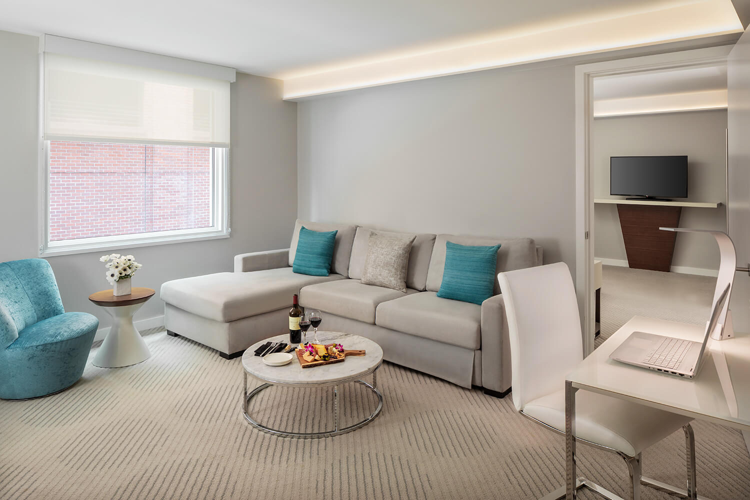 In a cozy, modern living room perfect for a Valentine's Month gathering, a beige sectional sofa adorned with turquoise pillows invites relaxation. A round coffee table sits nearby, while a flat-screen TV and a laptop on the white desk ensure entertainment and connectivity are always at hand.